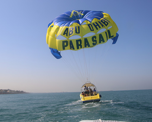 People enjoying a parasailing adventure over the turquoise waters of Abu Dhabi with a yellow and blue parachute labeled "Abu Dhabi Parasail".